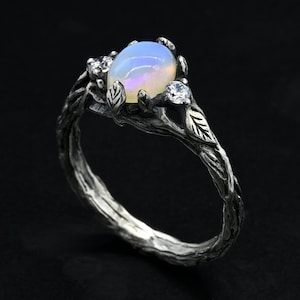 Könnte beinhalten: Ein silberner Ring mit einem Ast-Design, das mit einem weißen Opal-Edelstein und zwei kleineren klaren Edelsteinen besetzt ist. Der Ring ist auf einem schwarzen Hintergrund platziert.