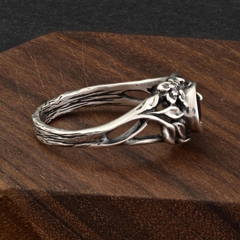 May include: A silver ring with a dark gemstone, featuring a nature-inspired design. The band has a textured, bark-like appearance, with floral and leaf accents around the gemstone setting. The ring is displayed on a wooden surface.