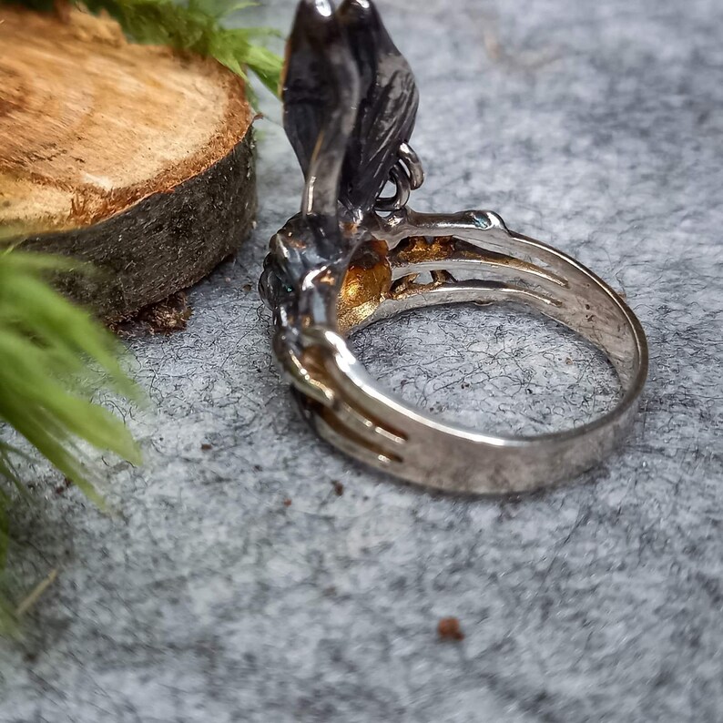 K&ouml;nnte beinhalten: Ein silberner Ring mit einem detaillierten Schmetterlingsdesign. Der Schmetterling hat eine dunkelgraue Oberfl&auml;che und ist mit einem gr&uuml;nen Edelstein besetzt. Der Ring liegt auf einer grauen, strukturierten Oberfl&auml;che.