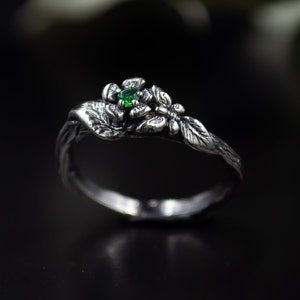 May include: A silver ring with a green gemstone set in a flower design. The ring is also decorated with a butterfly and leaves.