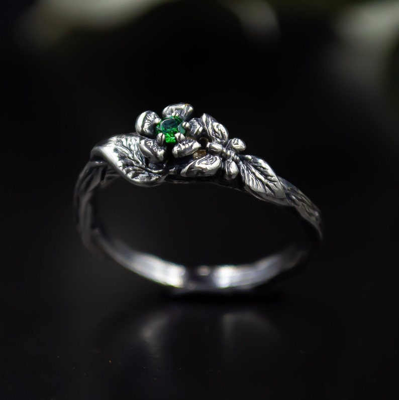 May include: A silver ring with a green gemstone set in a flower design. The ring is also decorated with a butterfly and leaves.