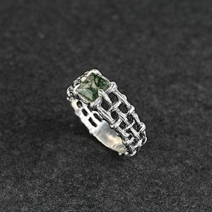 Op de afbeelding: Een zilveren ring met een vierkante, groene steen. De ring heeft een geweven ontwerp rond de band en de steen. De steen heeft een mosachtig uiterlijk. De ring staat tegen een donkere achtergrond.