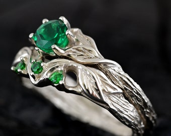 May include: A silver ring set with a large emerald green gemstone and smaller emerald green accent stones. The ring band is designed to look like a branch with leaves.