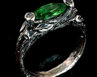 May include: A silver ring with a green gemstone and intricate details resembling a tree branch.
