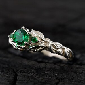 May include: A silver ring with a green gemstone and leaf details. The ring is set on a dark wooden surface.