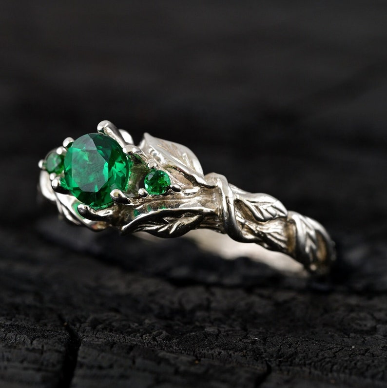 May include: A silver ring with a green gemstone and leaf details. The ring is set on a dark wooden surface.