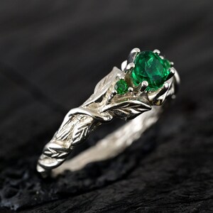 May include: A silver ring with a green emerald gemstone and leaf details. The ring is set on a dark wooden surface.