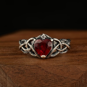 May include: A silver ring with a heart-shaped, faceted, red gemstone. The ring has a Celtic knot design around the heart and a small, clear stone at the top. The ring is displayed on a wooden surface.