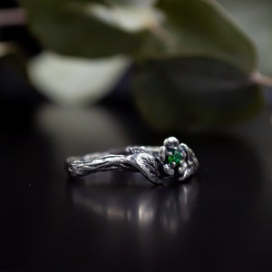 May include: A silver ring with a green gemstone set in a floral design. The ring band is textured to resemble a tree branch.