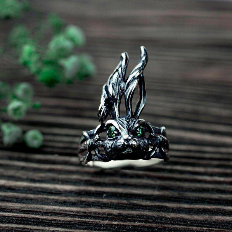 K&ouml;nnte beinhalten: Ein silberner Ring mit einem detaillierten Hasendesign. Der Hase hat gr&uuml;ne Augen und ist von einem Blattwerk umgeben.