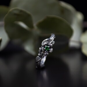 May include: A silver ring with a green gemstone set in a flower design. The ring is made to look like a delicate flower with leaves.