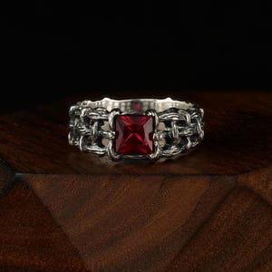 May include: A silver ring with a square, deep red gemstone at the center. The ring's band features a woven chain design. The ring is displayed on a dark wooden surface, creating a contrast with the bright gemstone.