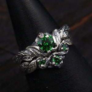 May include: A silver ring featuring a large green gemstone surrounded by smaller green gemstones. The ring is designed to resemble a branch with leaves.