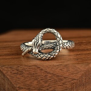 May include: A silver ring featuring a detailed snake design. The snake is coiled into a loop, with its head and tail meeting. The ring has a textured, scale-like pattern and is set against a wooden surface.