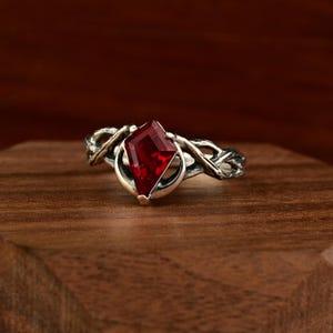 May include: A silver ring featuring a large, red, faceted gemstone. The ring's band has a twisted, vine-like pattern. The gemstone is a bright red and is set in a silver bezel.