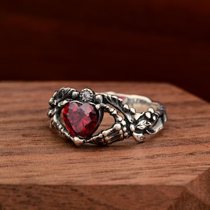 May include: A silver ring with a heart-shaped, faceted red gemstone. The ring features a detailed design with a skeleton hand holding the heart, accented with small leaves and a small clear stone. The ring is displayed on a wooden surface.