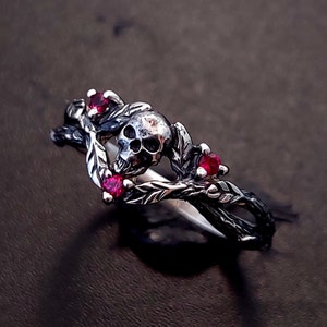 Puede incluir: Un anillo de plata con un dise&ntilde;o de calavera y piedras preciosas rojas. La calavera est&aacute; rodeada de hojas y ramas.