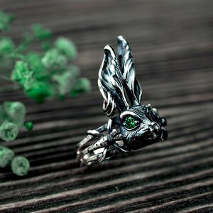 K&ouml;nnte beinhalten: Ein silberner Ring mit einem detaillierten Hasendesign. Die Augen des Hasen sind mit gr&uuml;nen Edelsteinen besetzt. Der Ring liegt auf einem Holzgrund.
