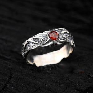 Puede incluir: Un anillo de plata con una piedra preciosa roja engastada en un diseño detallado y texturizado. El anillo está hecho de plata de ley y presenta un patrón único e intrincado.
