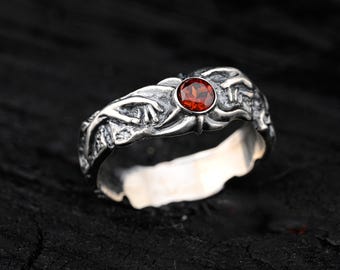 Puede incluir: Un anillo de plata con una piedra preciosa roja engastada en un diseño detallado y texturizado. El anillo está hecho de plata de ley y presenta un patrón único e intrincado.