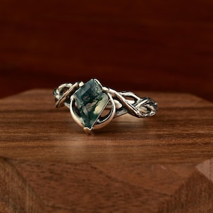 May include: A silver ring with a green moss agate gemstone in a diamond shape. The ring's band has a nature-inspired design with intertwined branches. The gemstone is set in a bezel setting, displayed on a wooden surface.