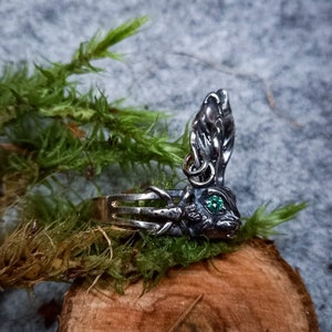 K&ouml;nnte beinhalten: Ein silberner Ring mit einem detaillierten Hasendesign. Der Hase hat einen gr&uuml;nen Edelstein als Auge und seine Ohren sind aufgestellt. Der Ring liegt auf einer Holzoberfl&auml;che mit gr&uuml;nem Moos.