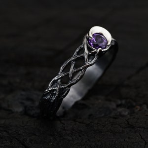 May include: A silver ring with a black oxidized finish features a delicate, intricate design resembling intertwined branches. A purple amethyst gemstone is set in the center, surrounded by a crescent moon shape.