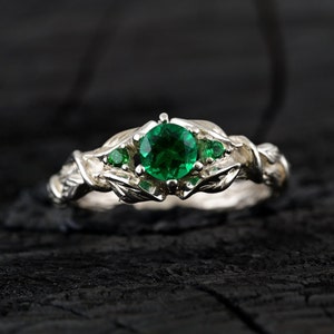 May include: A silver ring with a large green emerald gemstone surrounded by smaller green gemstones. The ring has a leafy design.