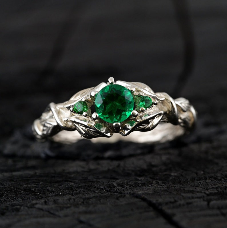 May include: A silver ring with a large green emerald gemstone surrounded by smaller green gemstones. The ring has a leafy design.