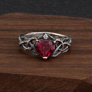 May include: A silver-toned ring with a heart-shaped, faceted, red gemstone. The ring features an intricate Celtic knot design around the gemstone and a small, clear accent stone above the heart. The ring is set on a dark wooden surface.