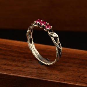 May include: A silver ring with a nature-inspired design. The ring features three vibrant red gemstones set in a twisted, branch-like band. A small leaf detail adds to the organic aesthetic. The ring is displayed on a wooden surface.