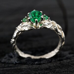 May include: A silver ring with a green emerald gemstone and smaller green gemstones on the band. The band is designed to look like a vine with leaves.