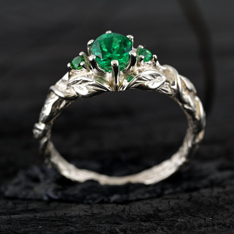 May include: A silver ring with a green emerald gemstone and smaller green gemstones on the band. The band is designed to look like a vine with leaves.