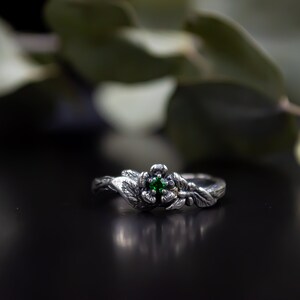May include: A silver ring with a green gemstone set in a flower design. The ring is surrounded by leaves and branches, creating a nature-inspired look.