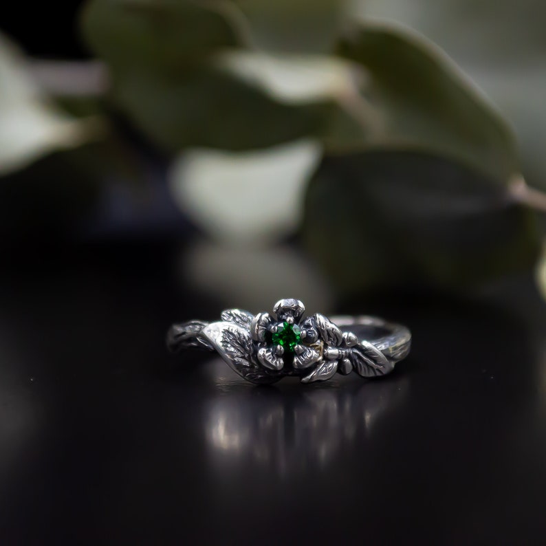 May include: A silver ring with a green gemstone set in a flower design. The ring is surrounded by leaves and branches, creating a nature-inspired look.