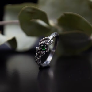 May include: A silver ring with a green gemstone set in a flower design. The ring is made to look like a branch with leaves.