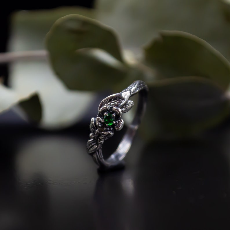 May include: A silver ring with a green gemstone set in a flower design. The ring is made to look like a branch with leaves.