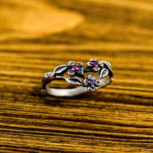 Könnte beinhalten: Ein silberner Ring mit einem floralen Design mit drei Blumen, die mit rosa und violetten Edelsteinen besetzt sind.