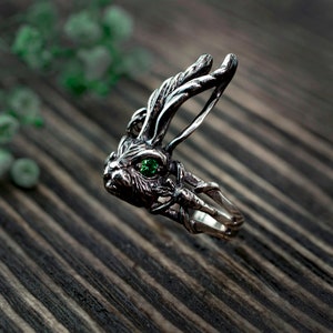 K&ouml;nnte beinhalten: Ein silberner Ring mit einem detaillierten Hasendesign und einem gr&uuml;nen Edelsteinauge. Der Ring liegt auf einer Holzoberfl&auml;che.
