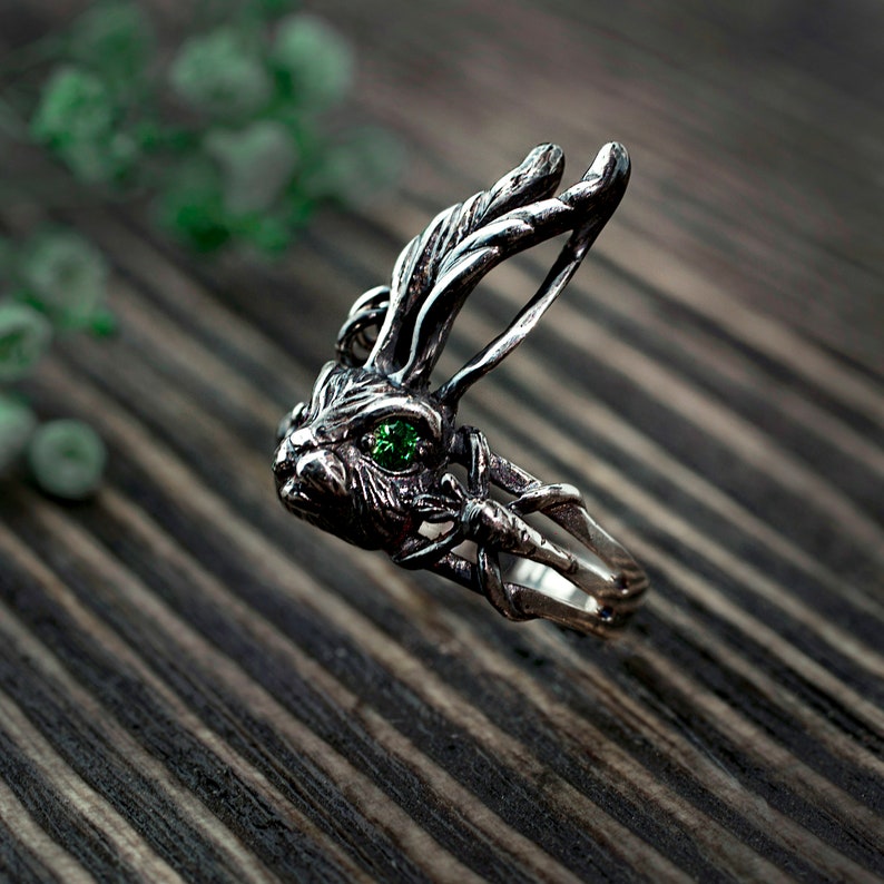 K&ouml;nnte beinhalten: Ein silberner Ring mit einem detaillierten Hasendesign und einem gr&uuml;nen Edelsteinauge. Der Ring liegt auf einer Holzoberfl&auml;che.