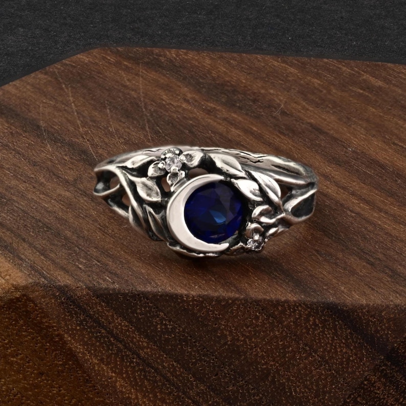 May include: A silver ring featuring a crescent moon design with a deep blue gemstone center. The ring is adorned with floral and leaf details, and small clear gemstones. The ring is set against a dark wooden surface.