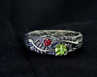 May include: A silver ring with a leafy design and two gemstones. The larger gemstone is a green peridot, and the smaller gemstone is a red garnet.