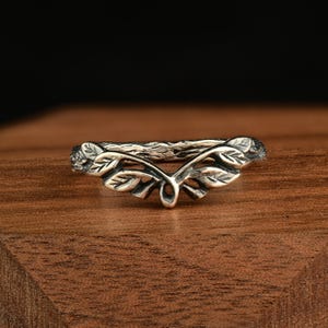 May include: A silver ring with a leaf design. The ring has a textured band, resembling bark, and detailed leaves arranged symmetrically. The design evokes a natural, botanical theme, with the leaves adding a delicate touch to the ring's overall appearance.