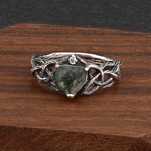 May include: A silver ring with a heart-shaped moss agate stone. The ring features an intricate Celtic knot design around the stone and band, with a small clear stone accent. The ring is set against a wooden background.