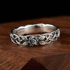 Puede incluir: Un anillo de plata con un diseño floral. El anillo presenta una rosa en el centro, rodeada de hojas y patrones entrelazados. El anillo está sobre un fondo oscuro.