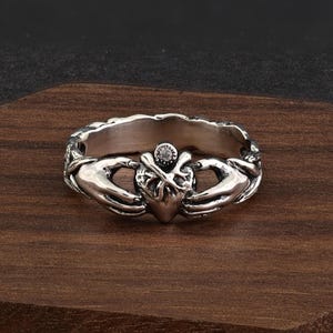 Könnte beinhalten: Silberner Claddagh-Ring mit Herz, Händen und Krone. Das Herz ist zentral, wird von zwei Händen gehalten und von einer kleinen Krone mit einem klaren Stein gekrönt. Auf dunklem Holzmaserungshintergrund.