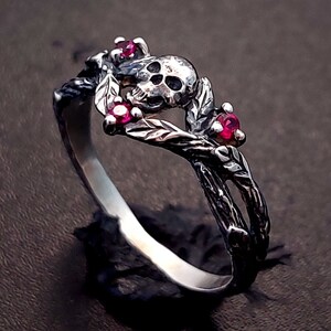 Puede incluir: Un anillo de plata con un dise&ntilde;o de calavera y piedras preciosas rojas. El anillo est&aacute; hecho de ramas y hojas retorcidas.