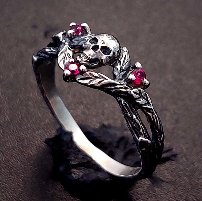 Puede incluir: Un anillo de plata con un dise&ntilde;o de calavera y piedras preciosas rojas. El anillo est&aacute; hecho de ramas y hojas retorcidas.