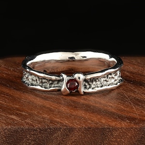 Può includere: Un anello d'argento con una fascia strutturata e una piccola gemma rossa rotonda. L'anello è posto su una superficie di legno, evidenziando il suo design dettagliato e il colore vibrante della pietra.