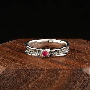 May include: A silver ring with a textured band and a small, round, red gemstone. The ring is set against a dark background, highlighting its intricate design and the vibrant color of the gemstone. The ring appears to be made of sterling silver.